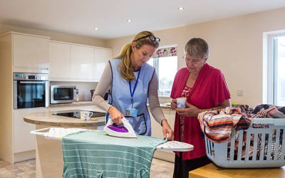 Care worker making a home visit. Female carer is ironing in the kitchen to help an elderly woman. The elderly woman is standing behind her chatting.